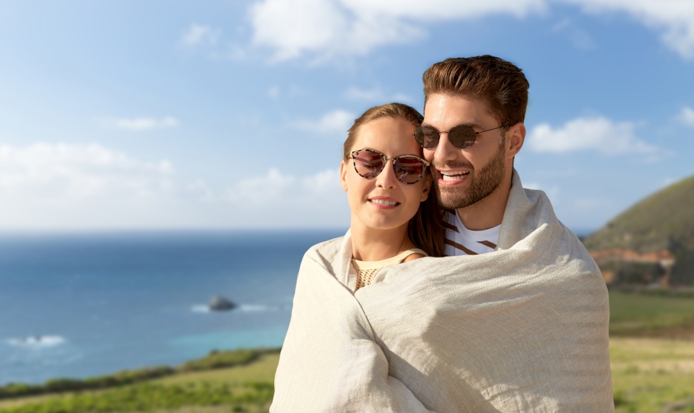 love, travel and tourism concept - happy couple in sunglasses covered with blanket hugging over big sur coast of california background. happy couple covered with blanket in california