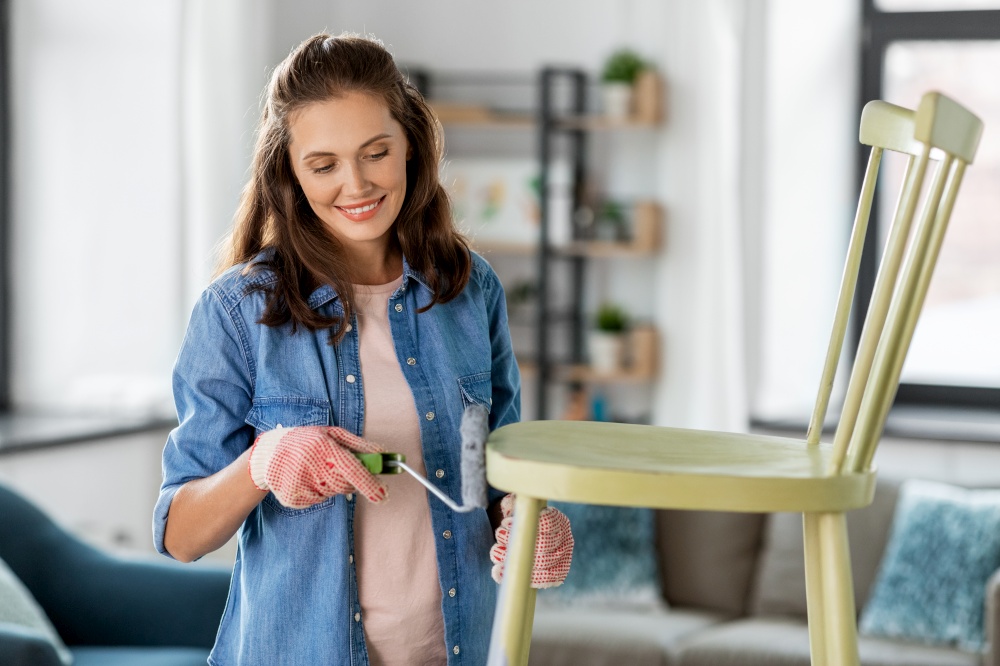 renovation, diy and home improvement concept - happy smiling woman in gloves with paint roller painting old wooden chair in grey color. woman painting old chair in grey color at home