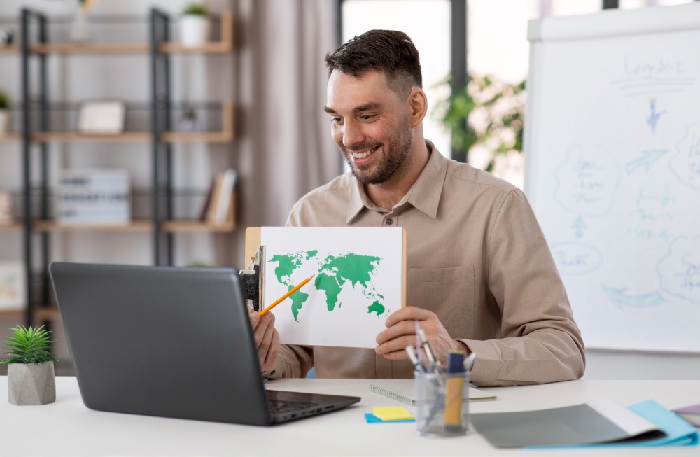 distant education, school and remote job concept - happy smiling male geography teacher with world map and laptop computer having online geography class at home office. teacher with world map having online class at home