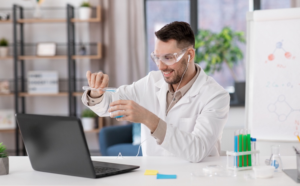 distant education, school and science concept - happy male chemistry teacher in goggles and earphones with laptop having online class and pouring chemical from test tube to flask at home office. chemistry teacher with laptop having online class