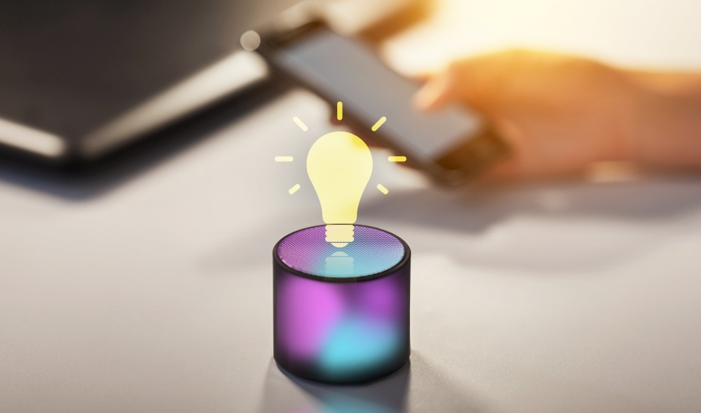 technology and ai concept - glowing smart speaker with virtual light bulb icon on table at office and virtual hologram of light bulb. smart speaker and virtual hologram of light bulb