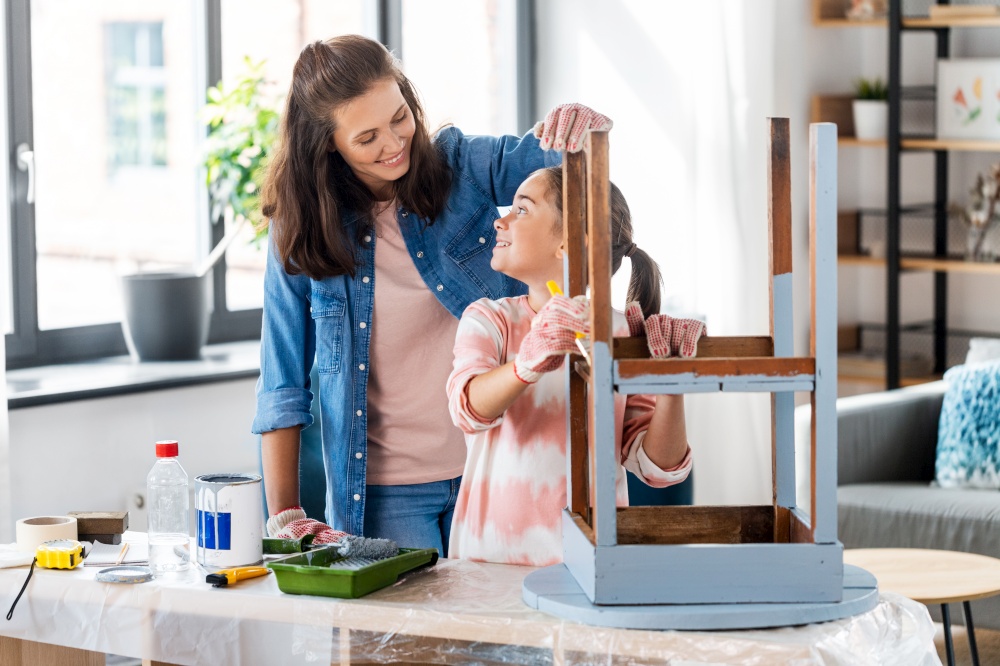 renovation, diy and home improvement concept - mother and daughter in gloves with paint brush painting old wooden table in grey color at home. mother and daughter painting old table at home