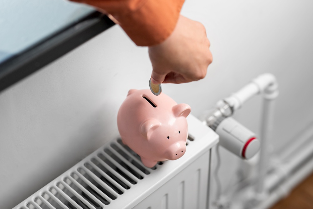 heating, energy crisis and consumption concept -close up of hand putting coin into piggy bank on radiator at home. hand putting coin into piggy bank on radiator