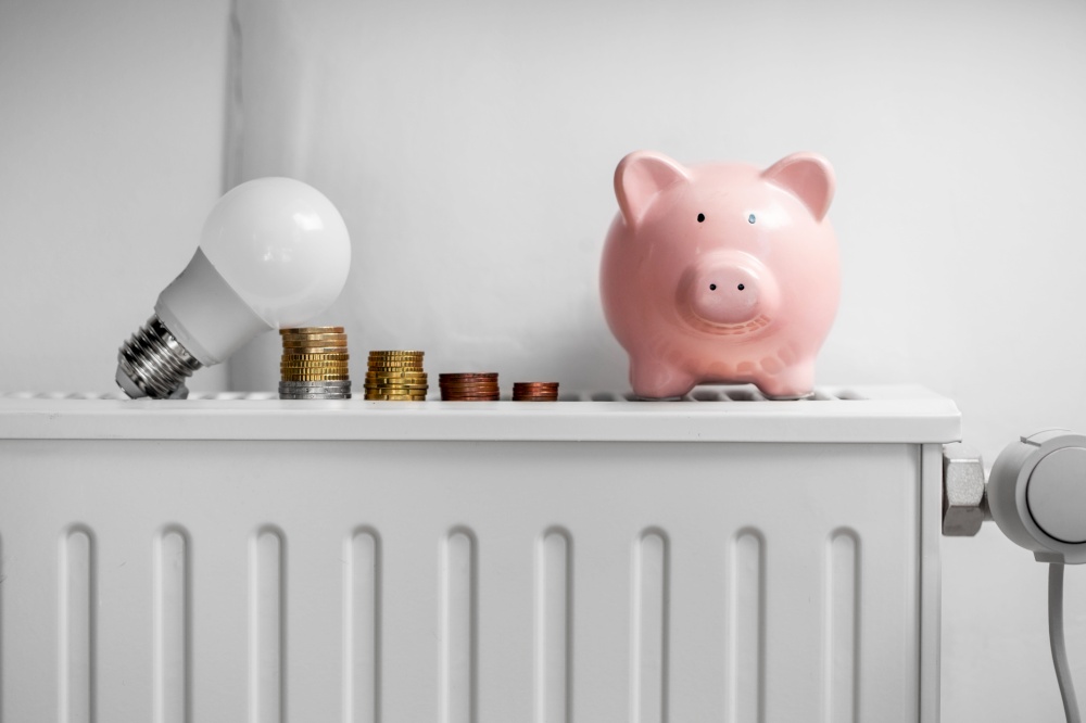 heating, energy crisis and consumption concept - piggy bank, light bulb and money on radiator at home. piggy bank, light bulb and coins on radiator