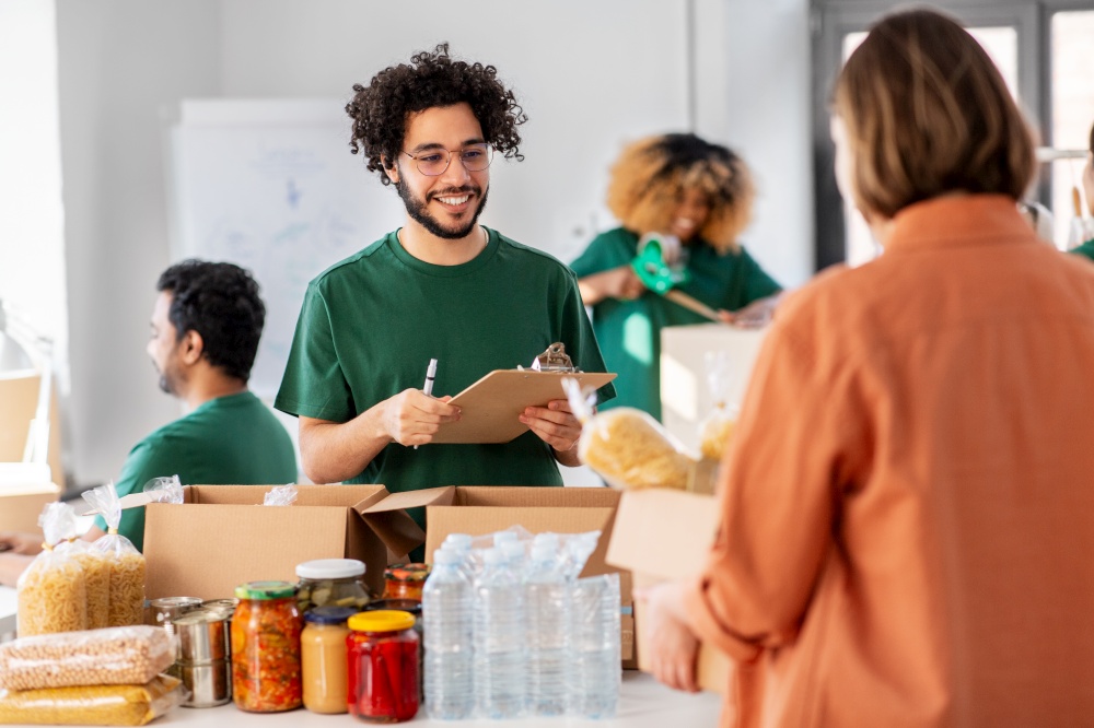 charity, donation and volunteering concept - happy smiling male volunteer with clipboard and woman taking box of food at distribution or refugee assistance center. happy volunteers packing food in donation boxes