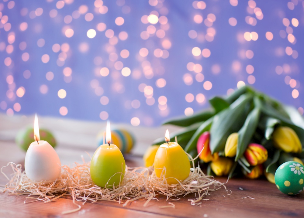 easter, holidays, tradition and object concept - burning candles in shape of eggs and tulip flowers over festive lights on violet background. candles in shape of easter eggs and tulip flowers