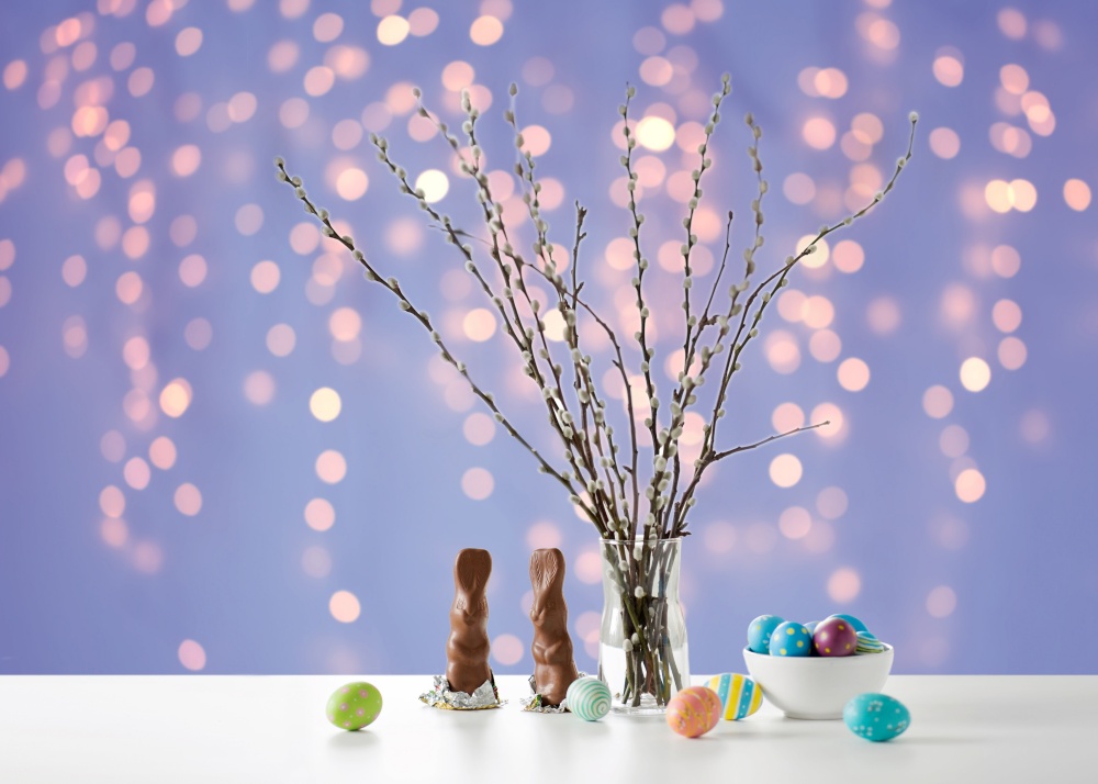 easter, holidays and object concept - pussy willow branches, colored eggs and chocolate bunnies on table over festive lights on violet background. pussy willow, easter eggs and chocolate bunnies