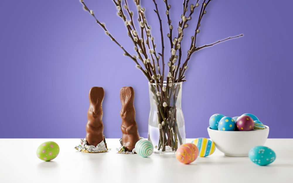 easter, holidays and object concept - pussy willow branches, colored eggs and chocolate bunnies on table over violet background. pussy willow, easter eggs and chocolate bunnies