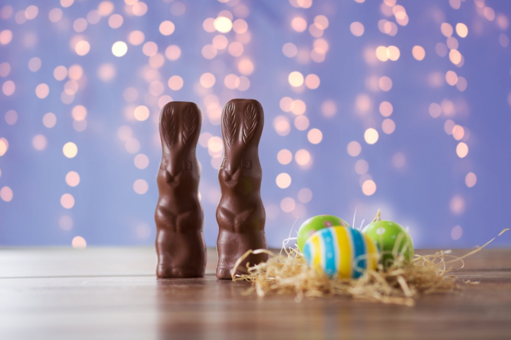 easter, holidays and object concept - close up of colored eggs in straw nest and chocolate bunnies on wooden table over festive lights on violet background. easter eggs in straw nest and chocolate bunnies
