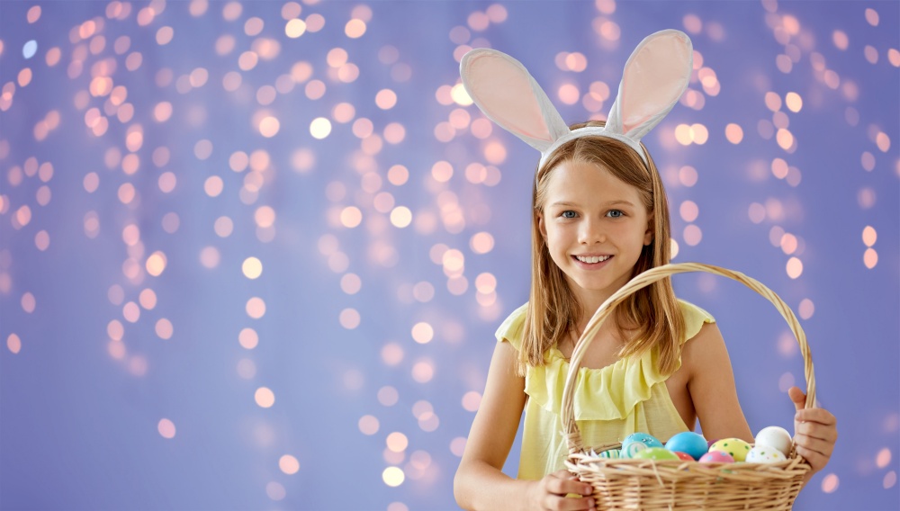 easter, holidays and people concept - happy girl wearing bunny ears headband with basket of colored eggs over festive lights on violet background. happy girl with colored easter eggs