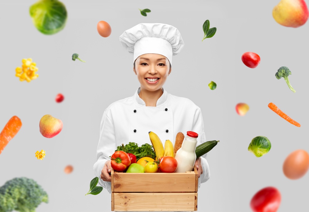 cooking, culinary and people concept - happy smiling female chef in toque holding food in wooden box over fruits and vegetables on grey background. happy smiling female chef with food in wooden box
