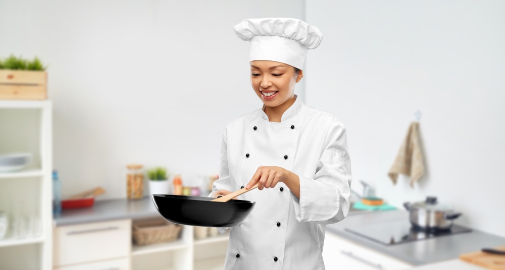food cooking, culinary and people concept - happy smiling female chef in toque with frying pan and wooden spatula over kitchen background. smiling female chef cooking food in frying pan