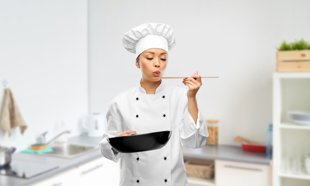 cooking, culinary and people concept - female chef with frying pan blowing to spatula and tasting food over kitchen background. female chef with frying pan tasting food