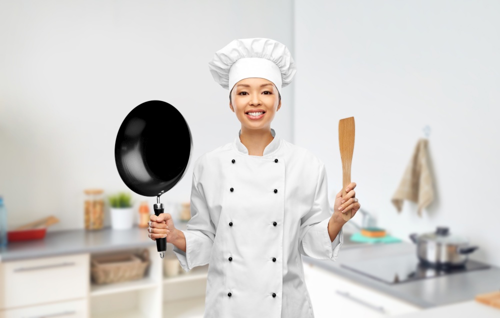 cooking, culinary and people concept - happy smiling female chef in toque with frying pan and spatula over kitchen background. smiling female chef with frying pan and spatula