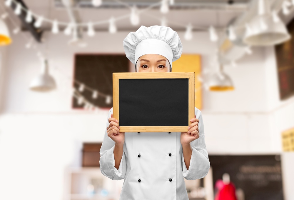 cooking, advertisement and people concept - female chef holding black chalkboard over restaurant background. female chef holding black chalkboard