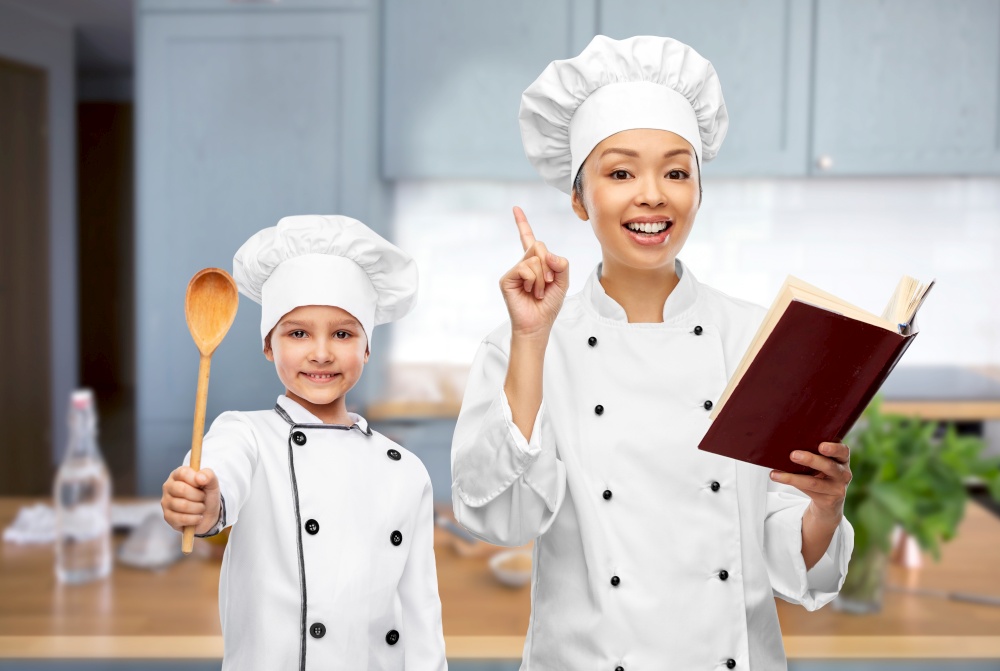 cooking, culinary and people concept - happy smiling female chef with cook book pointing finger up and little girl with wooden spoon over kitchen background. female chef with cook book and girl in kitchen