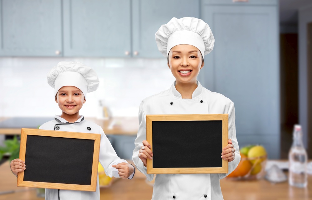 cooking, advertisement and people concept - happy smiling female chef and little girl holding black chalkboards over kitchen background. smiling female chef and girl holding chalkboards