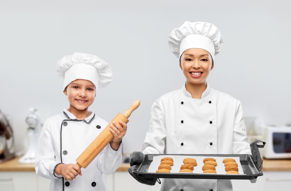 cooking, culinary and bakery concept - happy smiling female chef or baker in toque holding baking tray with oatmeal cookies and little girl with rolling pin over kitchen background. female chef and girl with cookies and rolling pin