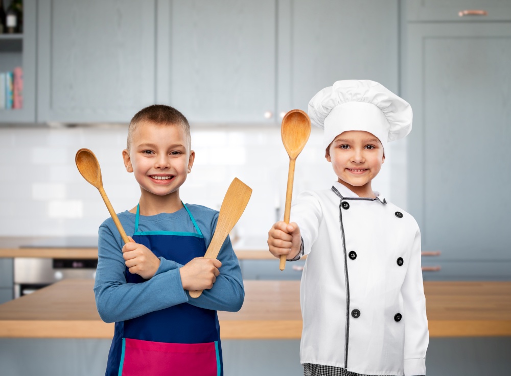 cooking, culinary and profession concept - happy smiling little boy in apron and girl in chef&rsquo;s toque with wooden spoon and spatula over kitchen background. happy children with spoon and spatula in kitchen