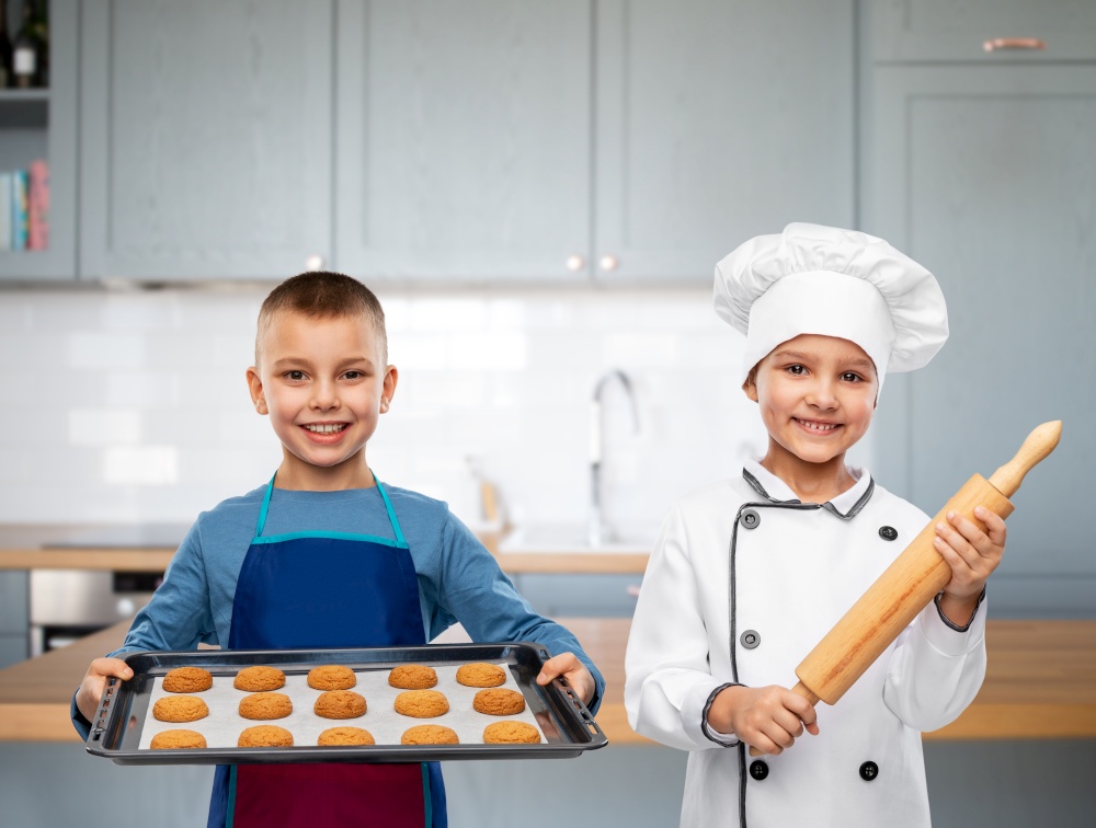 cooking, culinary and profession concept - happy smiling little girl in chef&rsquo;s toque with rolling pin and boy holding baking tray over kitchen background. children with rolling pin and cookies in kitchen