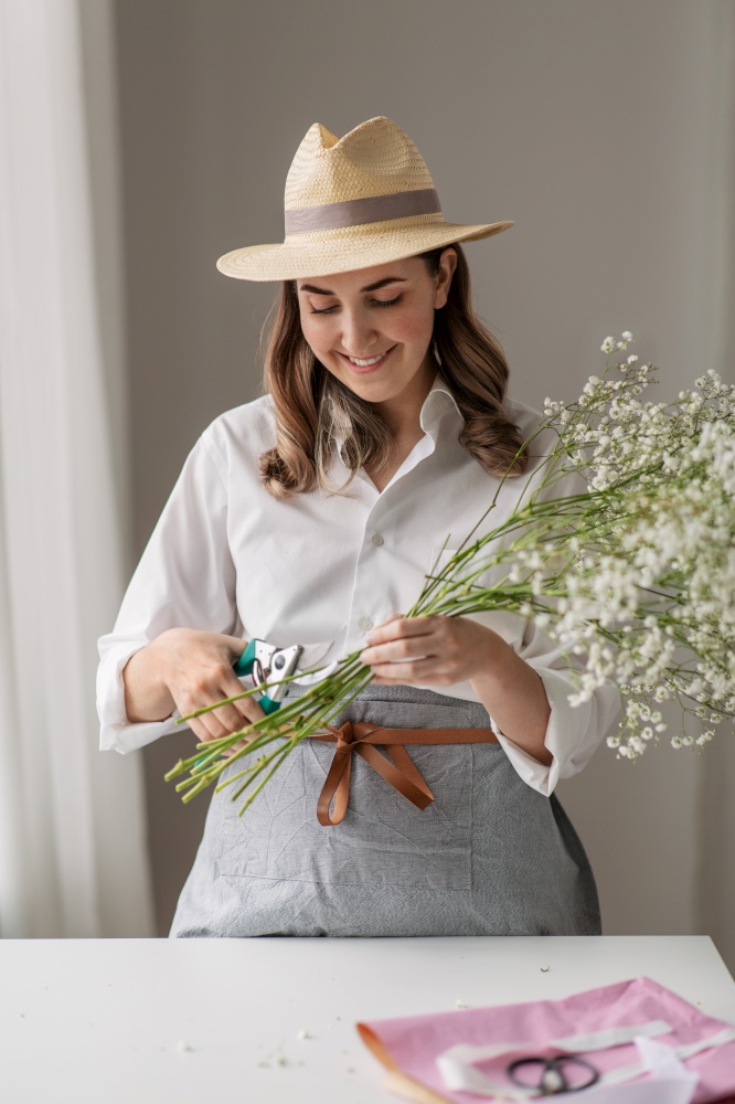 people, gardening and floral design concept - happy smiling woman or floral artist in straw hat making bunch of flowers and cutting gypsophila stems with pruning shears at home. happy woman making bunch of flowers at home