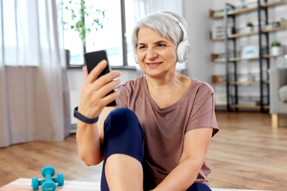 sport, fitness and healthy lifestyle concept - smiling senior woman with smartphone and wireless headphones exercising on mat at home. woman with phone and headphones exercising at home