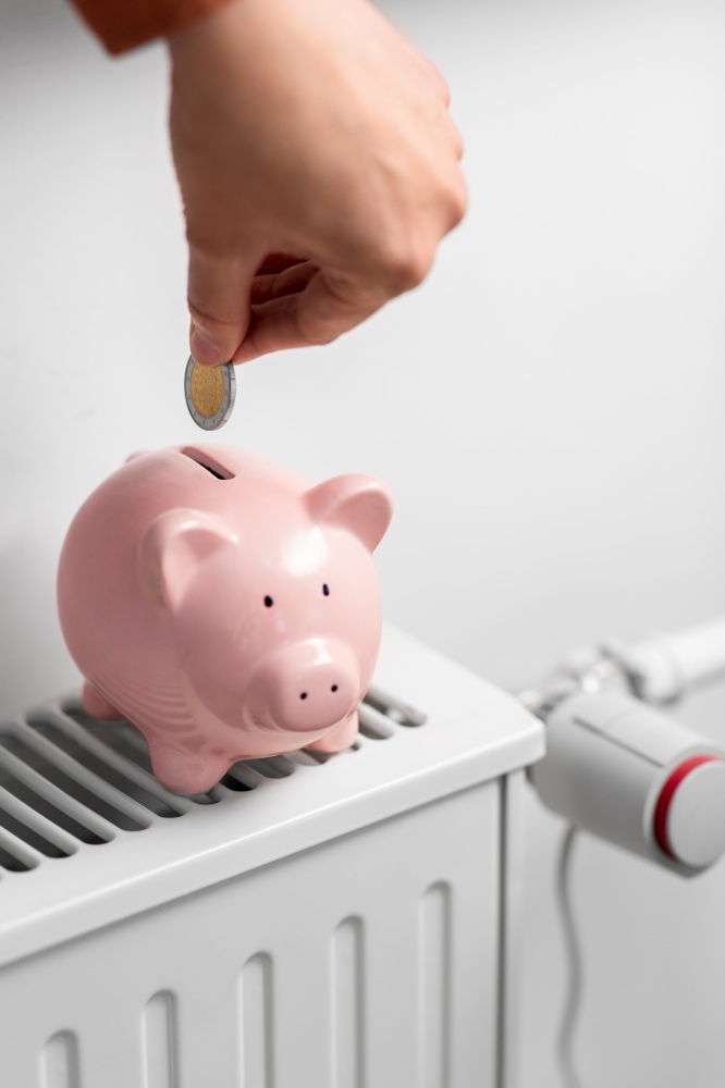 heating, energy crisis and consumption concept -close up of hand putting coin into piggy bank on radiator at home. hand putting coin into piggy bank on radiator