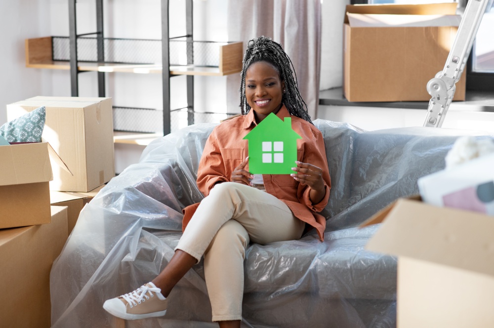 moving, people and real estate concept - happy smiling woman with green house and boxes sitting on sofa at new home. happy woman with green house and boxes moving home