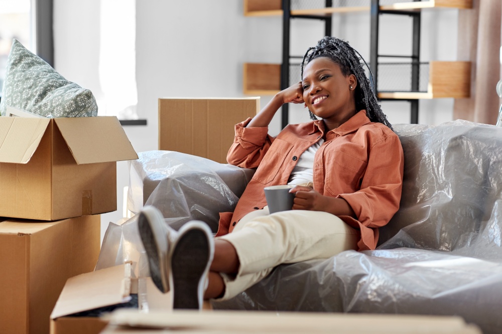 moving, people and real estate concept - happy smiling woman with boxes sitting on sofa with cup of coffee at new home. happy woman with boxes moving to new home