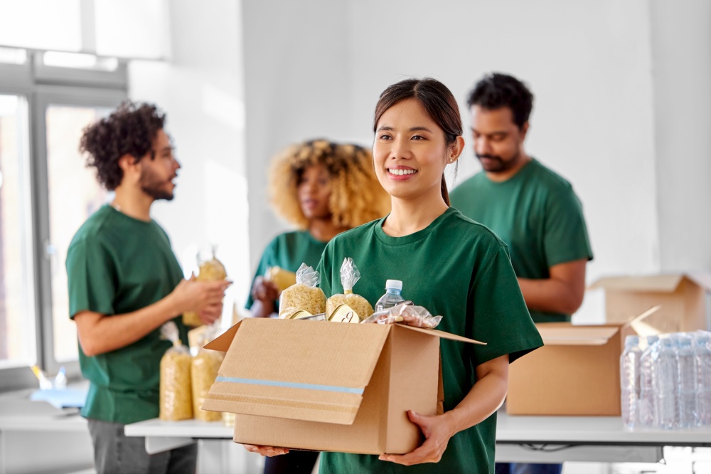 charity, donation and volunteering concept - happy smiling female volunteer with food in box and international group of people at distribution or refugee assistance center. happy volunteers packing food in donation boxes