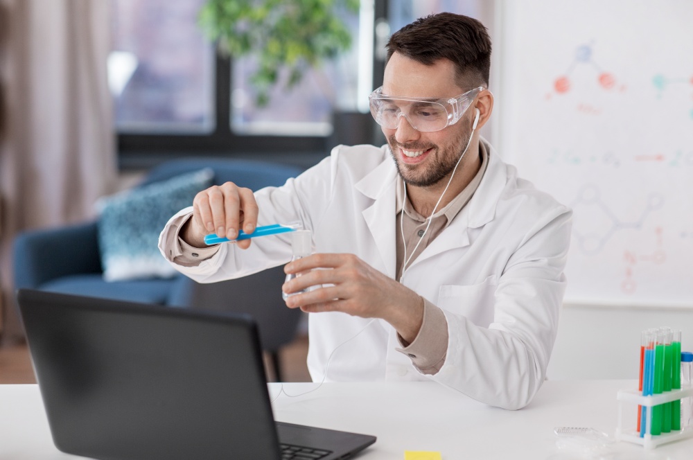 distance education, school and science concept - happy male chemistry teacher in goggles and earphones with laptop having online class and pouring chemical from test tube to flask at home office. chemistry teacher with laptop having online class