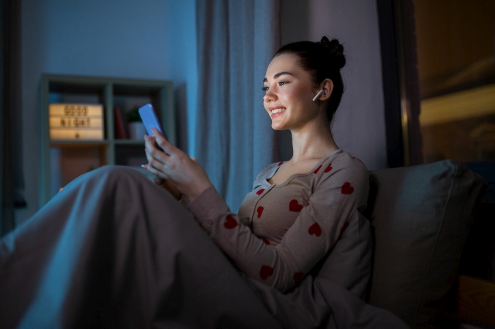 technology, bedtime and people concept - happy smiling teenage girl with smartphone and earphones sitting in bed at home at night. teenage girl with phone and earphones in bed at night