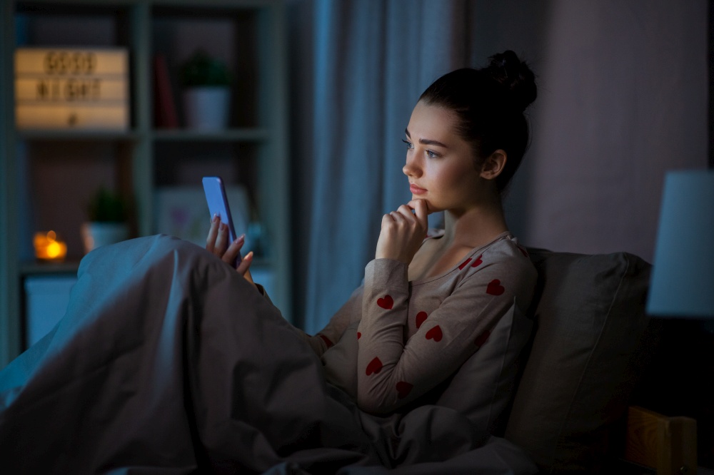 technology, bedtime and rest concept - pensive teenage girl in pajamas with smartphone sitting in bed at night. teenage girl in pajamas with phone in bed at night