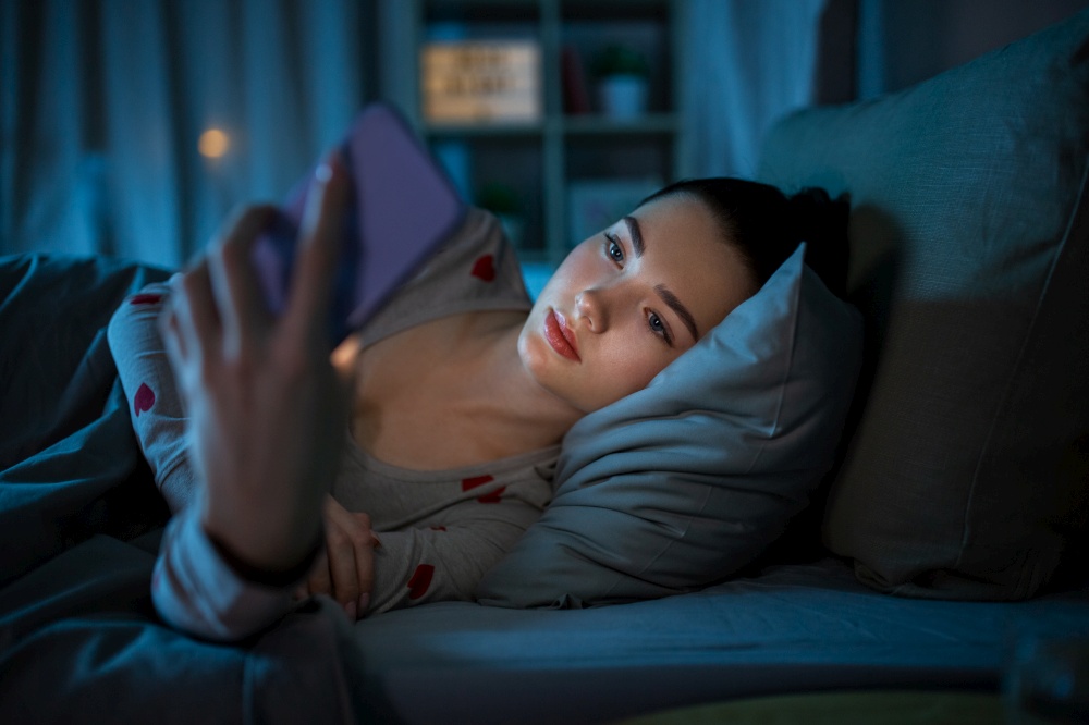 technology, bedtime and rest concept - teenage girl in pajamas with smartphone lying in bed at night. teenage girl with phone lying in bed at night