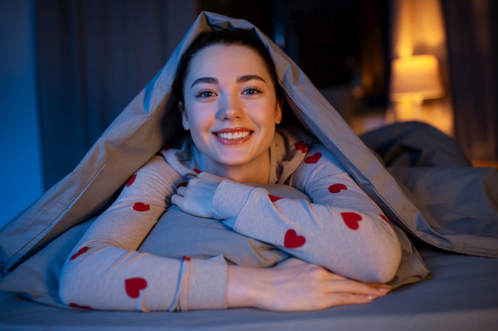 people, bedtime and rest concept - happy teenage girl lying under blanket in bed at home at night. happy teenage girl lying under blanket in bed
