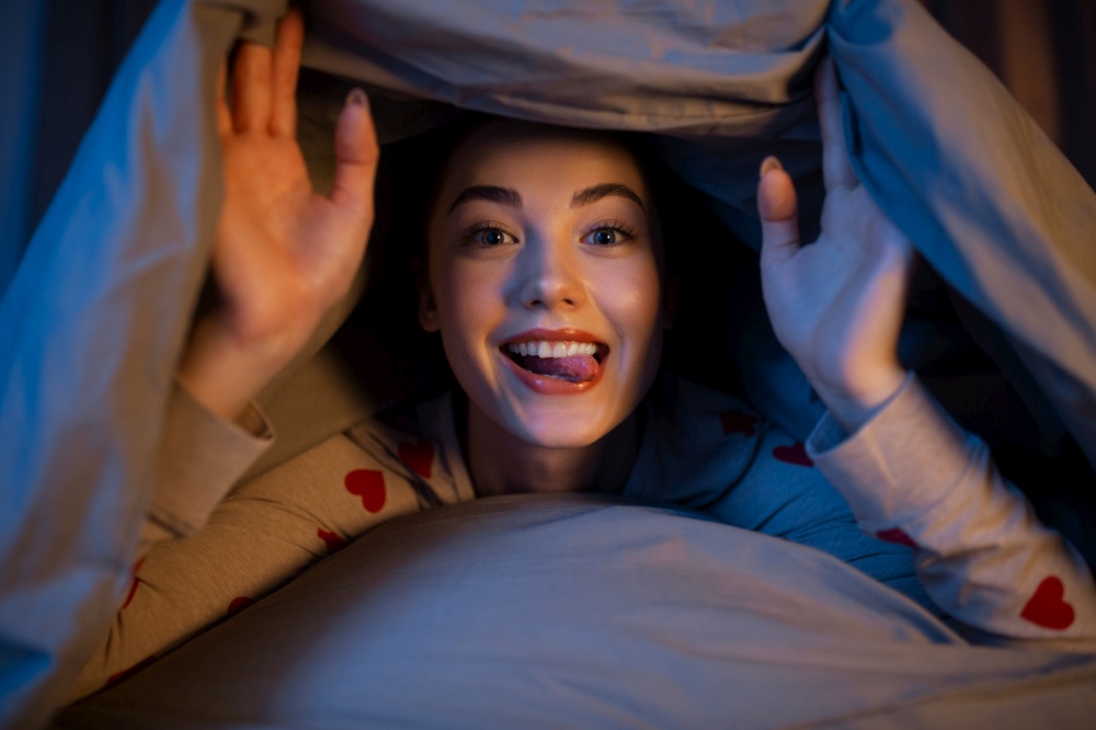 people, bedtime and rest concept - happy teenage girl lying under blanket in bed with her tongue stuck out at home at night. happy teenage girl lying under blanket in bed