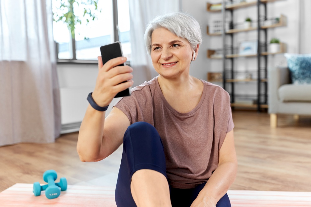 sport, fitness and healthy lifestyle concept - smiling senior woman with smartphone and wireless earphones exercising on mat at home. woman with phone and earphones exercising at home