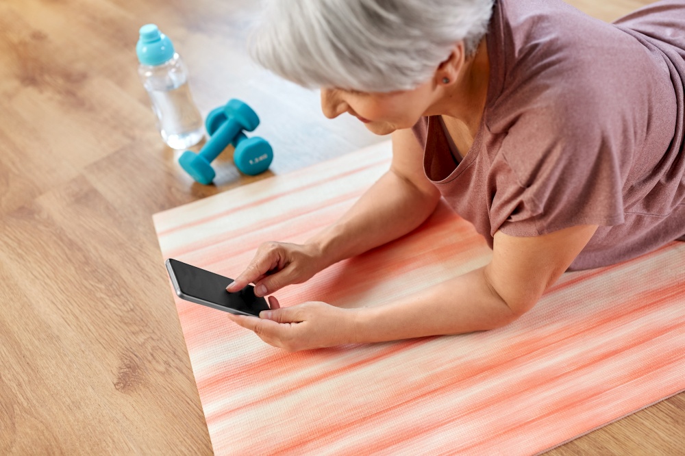 sport, fitness and healthy lifestyle concept - smiling senior woman with smartphone exercising on mat at home. senior woman with smartphone exercising at home