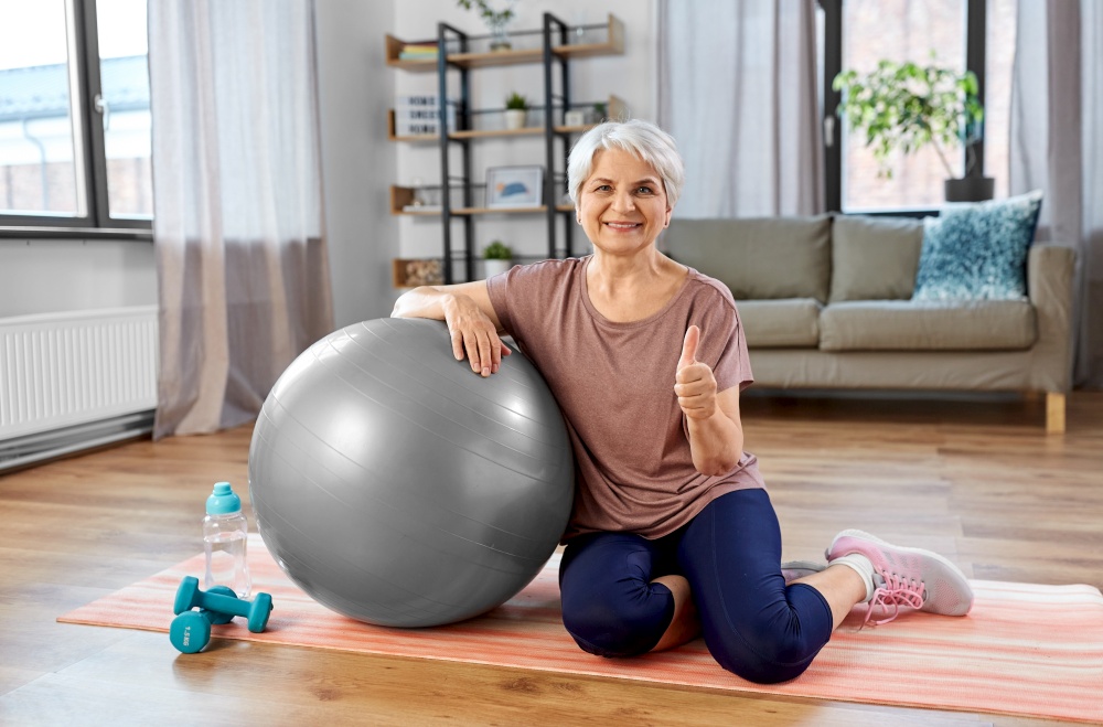 sport, fitness and healthy lifestyle concept - smiling senior woman with exercise ball showing thumbs up sitting on mat at home. smiling senior woman with exercise ball at home