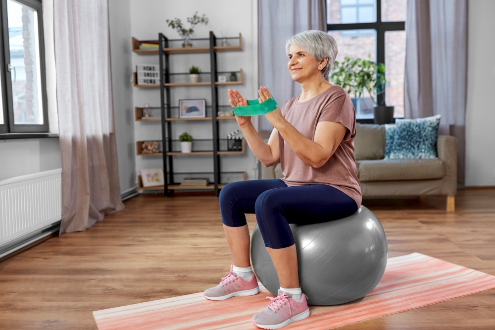 sport, fitness and healthy lifestyle concept - smiling senior woman with resistance band sitting on exercise ball at home. senior woman exercising with elastic band at home