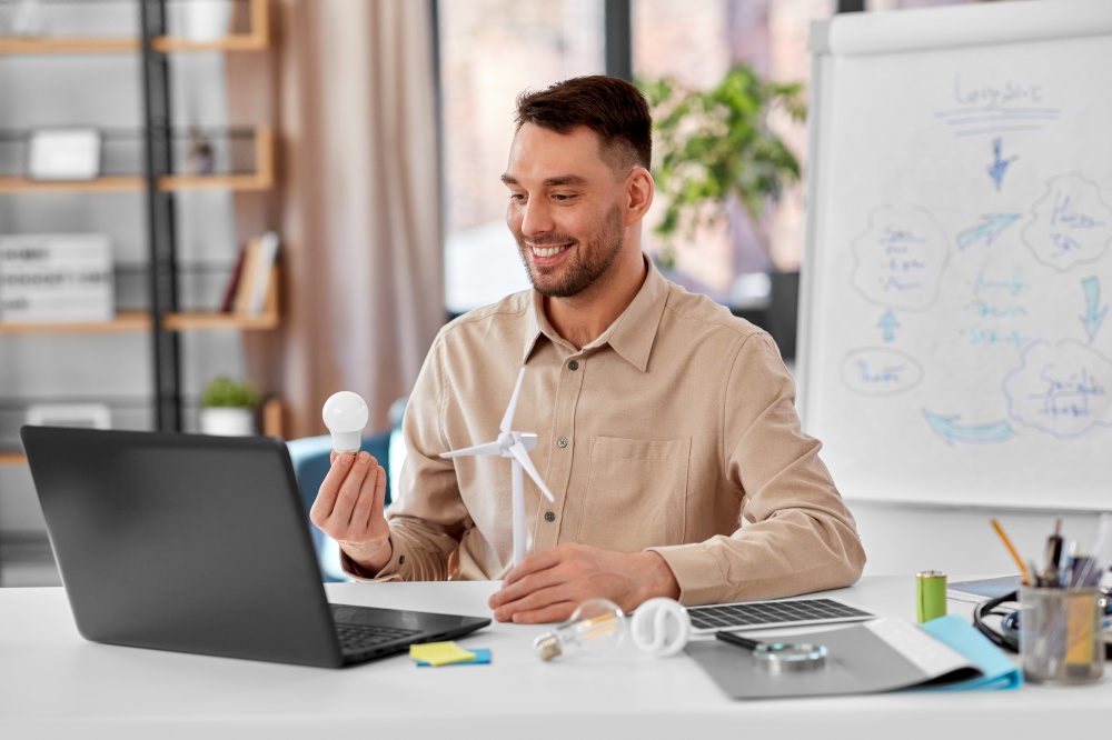 distance education, school and green energy concept - happy smiling male teacher with laptop computer, electric light bulb and wind turbine model having online class at home office. male teacher with laptop and wind turbine at home