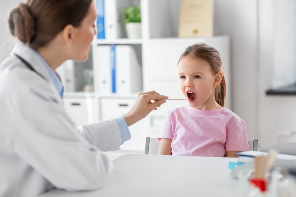 medicine, healthcare and pediatry concept - female doctor or pediatrician with tongue depressor checking little girl patient&rsquo;s throat on medical exam at clinic. female doctor and little girl patient at clinic