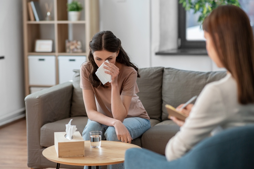 psychology, mental health and people concept - crying woman patient wiping tears with paper tissue and psychologist at psychotherapy session. crying woman and psychologist at psychotherapy