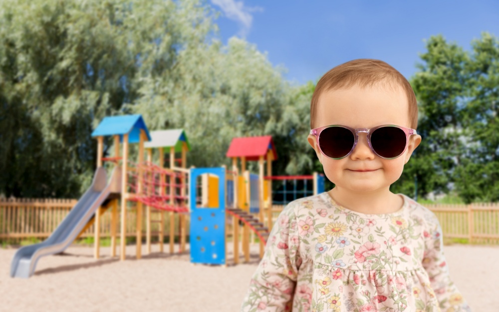 childhood, summer and people concept - happy little baby girl in sunglasses over children&rsquo;s playground background. happy baby girl in sunglasses over playground