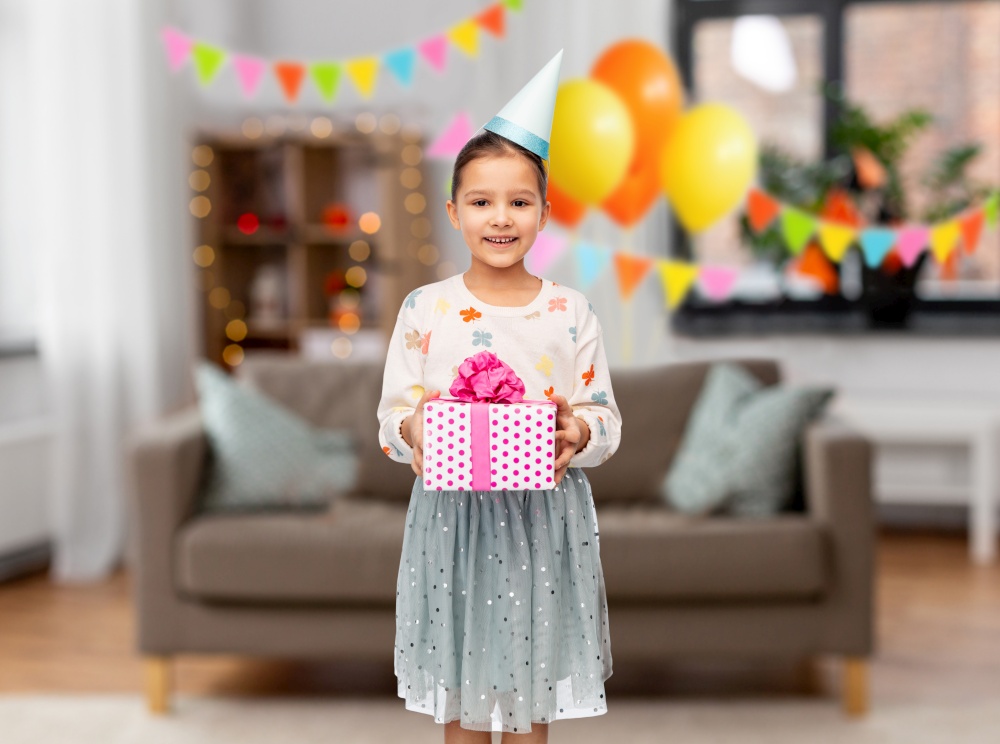 birthday, childhood and people concept - portrait of smiling little girl in party hat with gift box decorated living room background. smiling girl in party hat with birthday gift