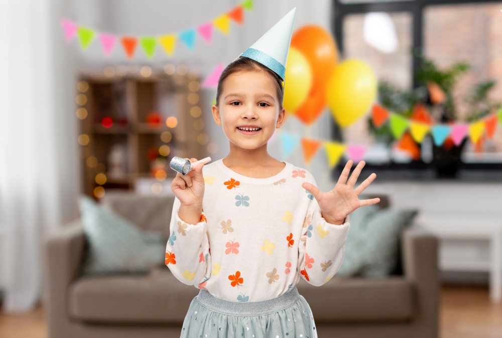 birthday, childhood and people concept - portrait of smiling little girl in party hat with blower having fun over decorated living room background. smiling girl in birthday party hat with blower