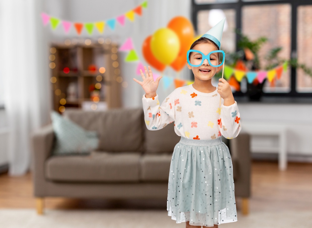 birthday, childhood and people concept - portrait of little girl in party hat with glasses over decorated living room background. little girl in birthday party hat with glasses