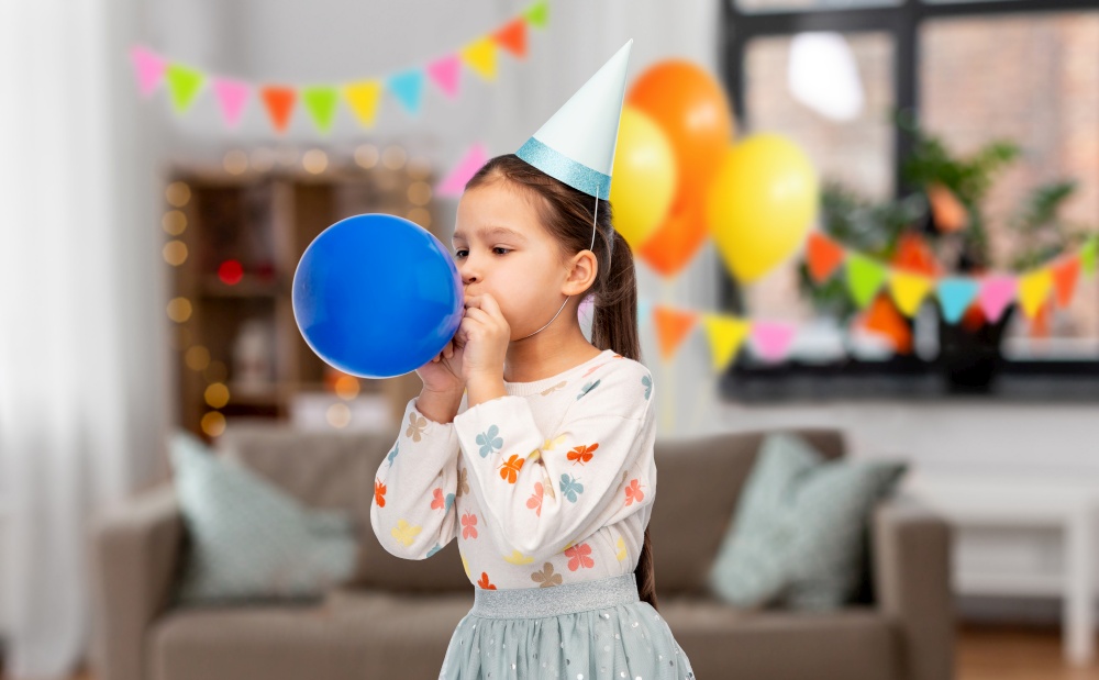 birthday, childhood and people concept - portrait of little girl in party hat blowing balloon over decorated living room background. little girl in birthday party hat blowing balloon