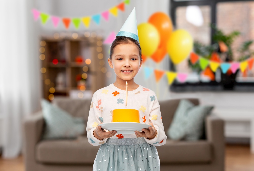 birthday, childhood and people concept - portrait of smiling little girl in party hat with cake over decorated living room background. smiling girl in party hat with birthday cake