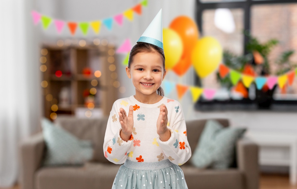 birthday, childhood and people concept - portrait of smiling little girl in party hat applauding over. happy little girl in birthday party hat applauding